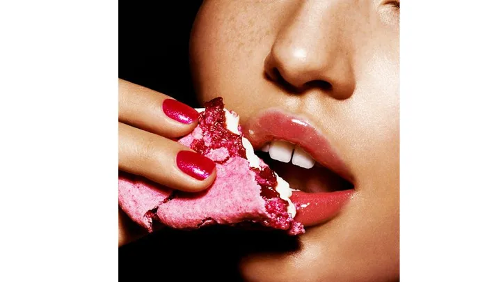 Close-up of a woman's face with pink lips, eating a pink and white macaron. Her nails are painted pink.