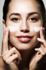 A woman applying white facial cream to her cheeks, smiling and looking at the camera.