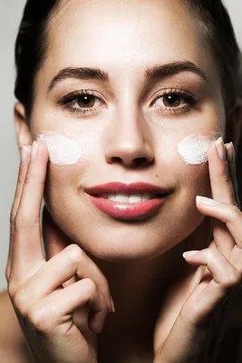 A woman applying white facial cream to her cheeks, smiling and looking at the camera.