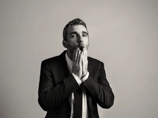 Alt text: "A man in a suit looks surprised with hands near his mouth in a black and white photograph."