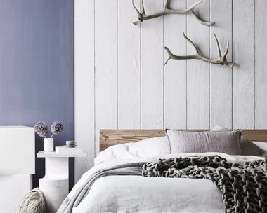 Modern minimalist bedroom with wooden bed, white bedding, knitted throw, antler wall decor, and side table with flowers.