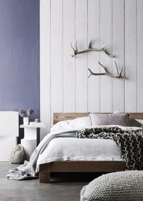 Modern minimalist bedroom with wooden bed, white bedding, knitted throw, antler wall decor, and side table with flowers.