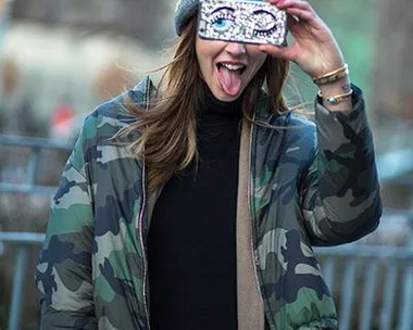 Person in camo jacket and beanie taking selfie with smartphone case featuring cartoon eyes, tongue out, playful pose.