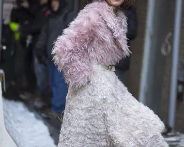 A woman in a stylish pink fur coat and white skirt walks on a snowy street, smiling confidently.