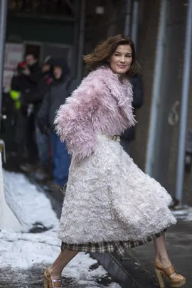 A woman in a stylish pink fur coat and white skirt walks on a snowy street, smiling confidently.