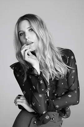 Woman in a sheer floral blouse and pants poses thoughtfully with hand near mouth in black and white photo.