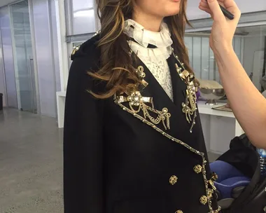 Model in ornate black jacket getting makeup touched up backstage at a photoshoot.