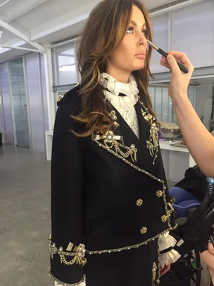 Model in ornate black jacket getting makeup touched up backstage at a photoshoot.