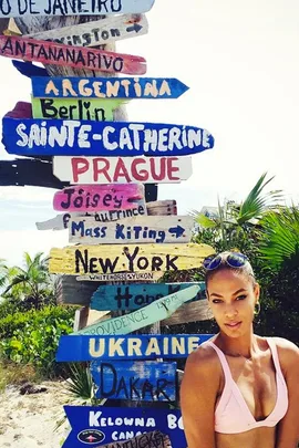 A woman in a pink bikini stands by a colorful international signpost on a sunny day, with palm trees in the background.