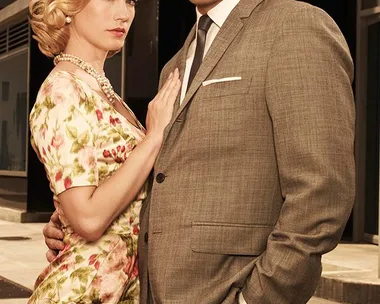 A man in a grey suit and a woman in a floral dress stand close together in a 1960s style scene.