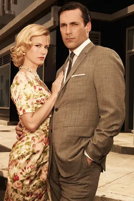 A man in a grey suit and a woman in a floral dress stand close together in a 1960s style scene.