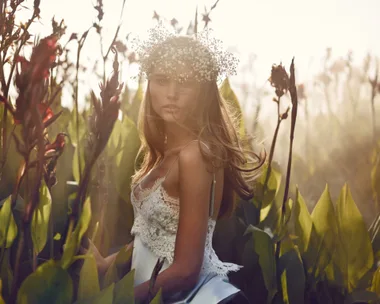 A woman in a white lace dress and flower crown stands in a field of tall plants, with sunlight illuminating her.