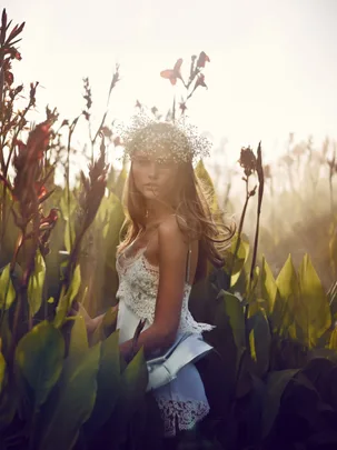 A woman in a white lace dress and flower crown stands in a field of tall plants, with sunlight illuminating her.