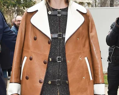 A woman in a stylish brown coat with white lapels and sunglasses stands outdoors, trees and people in the background.