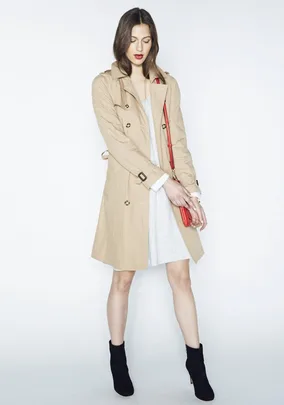 A woman in a beige trench coat, gray dress, black ankle boots, and red handbag poses against a plain white background.