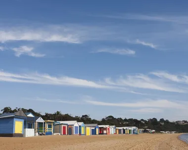 Colorful beach huts line up on a sandy shore under a blue sky with light clouds.