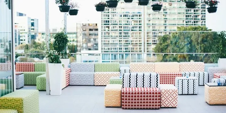 Colorful, patterned seating on an outdoor terrace of a hotel with city buildings and greenery in the background.