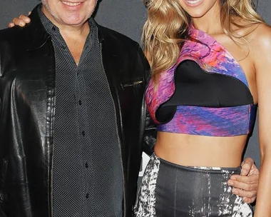 A man in a leather jacket and a woman in a colorful crop top and patterned skirt smile at a red carpet event.