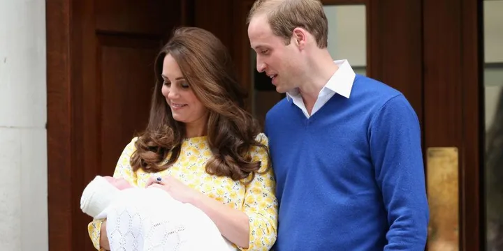 A couple smiles at their newborn baby in a white blanket outside a building. The woman wears a yellow dress; the man, a blue sweater.