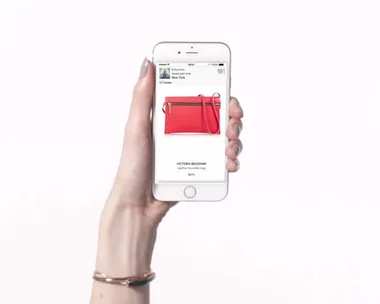 A hand holding a smartphone displaying a red Victoria Beckham leather shoulder bag.