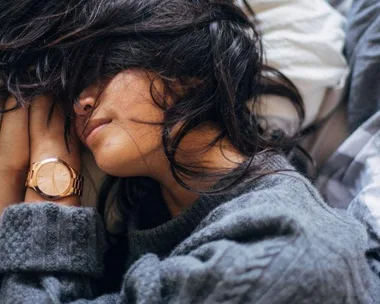 A woman with long hair, wearing a gray sweater, lies asleep in bed with her face partially covered.