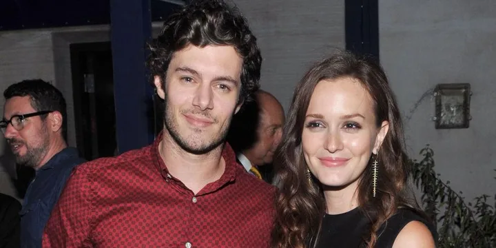 A smiling couple poses closely together; one wears a red shirt, the other a black dress with long hair and earrings.