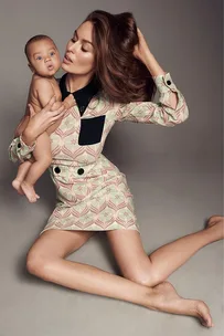 A woman in a patterned dress holds a baby in her lap while seated on the floor, posing with her hair.