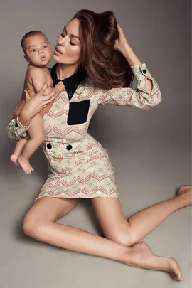 A woman in a patterned dress holds a baby in her lap while seated on the floor, posing with her hair.