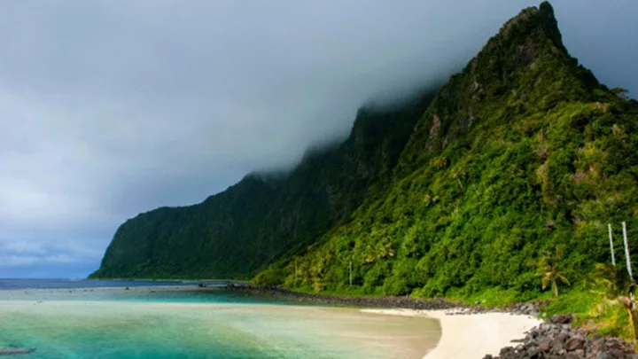 Beach view in Samoa with turquoise water, a sandy shore, and lush green mountains partially covered by clouds.