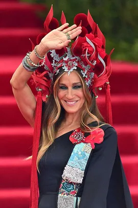 Person wearing a black dress, colorful sash, and red ornate headpiece, smiling and waving on a red carpet.