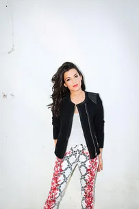 Woman with long dark hair in a black jacket and white floral pants standing against a white wall.