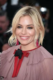 Sienna Miller at Cannes Film Festival, wearing a pink dress with red accents and red earrings, smiling at the camera.