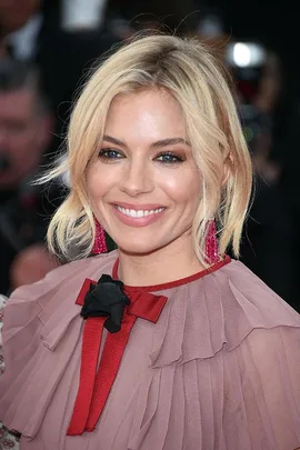 Sienna Miller at Cannes Film Festival, wearing a pink dress with red accents and red earrings, smiling at the camera.