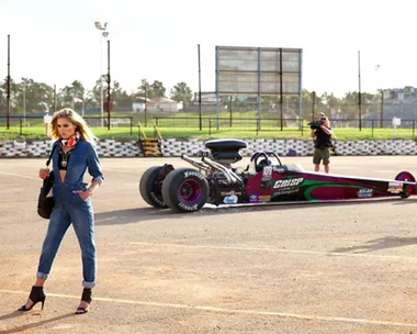 Model in denim outfit poses at racetrack near drag racing car; cameraman in background.