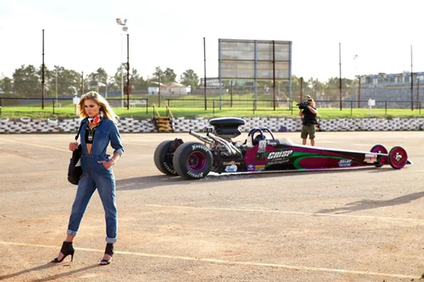 Model in denim outfit poses at racetrack near drag racing car; cameraman in background.