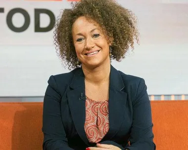 Rachel Dolezal on the Today show set, wearing a navy blazer and patterned blouse, with a smile, seated on an orange couch.