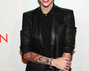 Person with short hair, tattoos, and a black outfit smiling at an event with a "Women" backdrop.