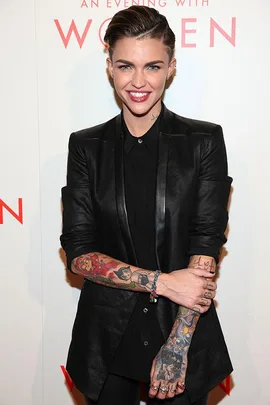 Person with short hair, tattoos, and a black outfit smiling at an event with a "Women" backdrop.