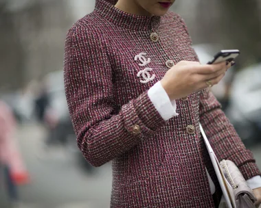 A person in a Chanel coat uses a smartphone while carrying a beige clutch.