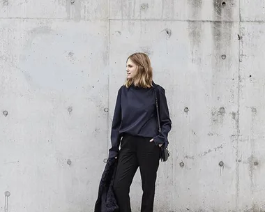 A woman in dark casual clothes stands against a concrete wall, holding a coat and wearing white sneakers.