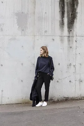 A woman in dark casual clothes stands against a concrete wall, holding a coat and wearing white sneakers.