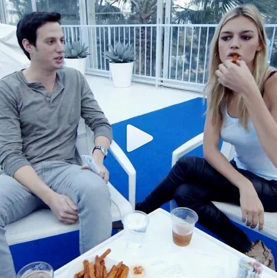 A man and a woman are sitting on chairs eating food from a table at an outdoor setting.