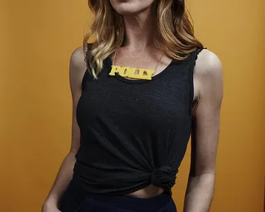 A woman with shoulder-length hair wearing a black tank top and a yellow necklace that says "POLLY," against an orange background.