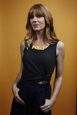 A woman with shoulder-length hair wearing a black tank top and a yellow necklace that says "POLLY," against an orange background.