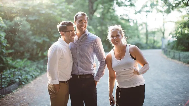 A couple posing for an engagement photo in a park is photobombed by a woman in workout clothes.