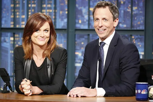 Amy Poehler and Seth Meyers sitting at a desk, smiling on the set of a talk show with city lights in the background.