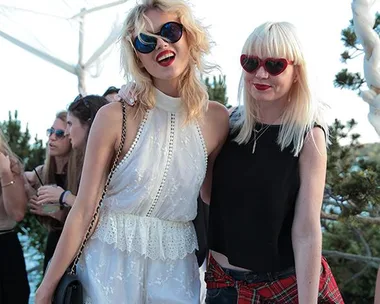 Two women in stylish outfits and sunglasses, smiling at a sunny outdoor party with people in the background.