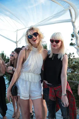 Two women in stylish outfits and sunglasses, smiling at a sunny outdoor party with people in the background.