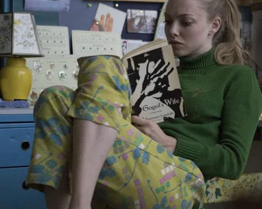 A woman in casual clothing sits barefoot in a colorful room, reading a book titled "Gogol's Wife".