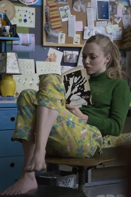 A woman in casual clothing sits barefoot in a colorful room, reading a book titled "Gogol's Wife".